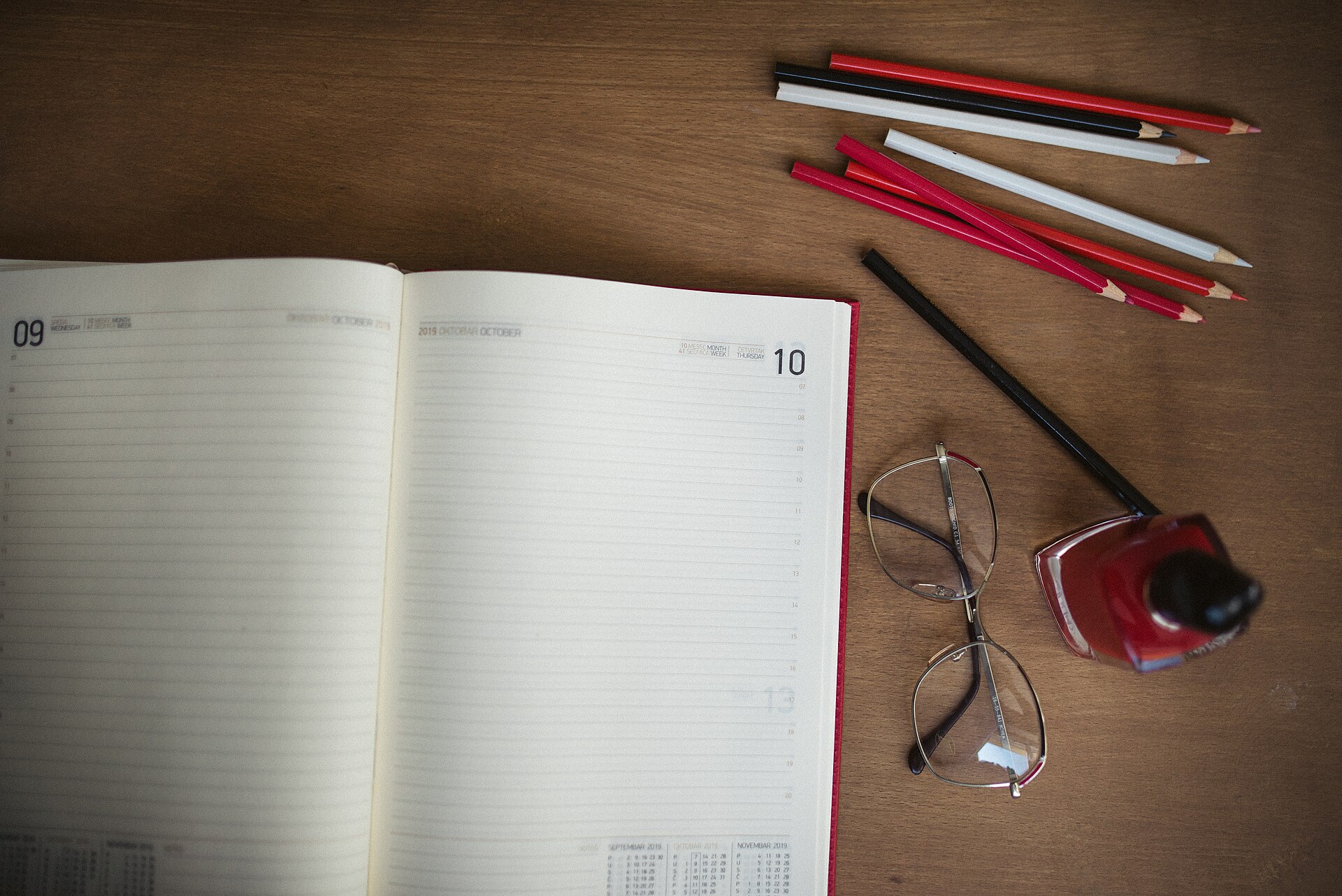 An open planner with blank weekly pages beside coloured pencils and glasses on a wooden desk